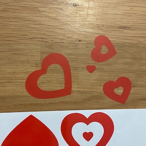 May include: Several red heart-shaped stickers of varying sizes are arranged on a wooden surface. Some hearts have a heart-shaped cutout in the center. The stickers are likely for crafts or decorations.