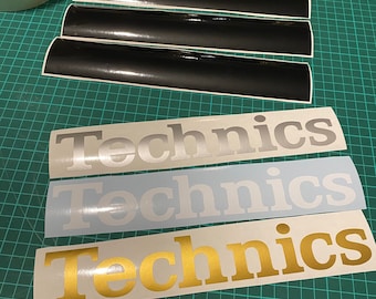 Może przedstawiać: Trzy naklejki winylowe z napisem "Technics" w różnych kolorach. Górna naklejka jest srebrna, środkowa naklejka jest biała, a dolna naklejka jest złota.