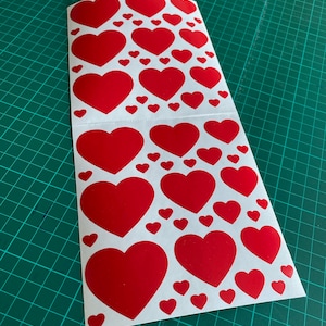 May include: A sheet of white paper with multiple red heart stickers of various sizes. The stickers are arranged in a pattern.
