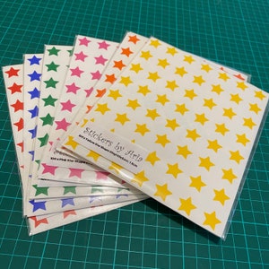 May include: A set of six sheets of star-shaped vinyl stickers. Each sheet contains 100 yellow, pink, red, blue, green, and orange star stickers. The stickers are 1.5 cm in size. The text on the sheet reads "Stickers by Aria 100 x Yellow Star Shape Vinyl Stickers 1.5cm 100 x Pink Star Shape"
