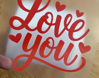 Może przedstawiać: Przezroczysta naklejka z napisem "Love you" w czerwonym skrypcie, z małymi serduszkami. Naklejka znajduje się na jasnej powierzchni, odpowiednia do rękodzieła i prezentów.