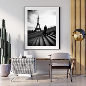 Può includere: Stampa incorniciata in bianco e nero della Torre Eiffel a Parigi. L'immagine mostra una coppia su una scala, con la famosa torre sullo sfondo. La stampa è esposta in una stanza con scrivania, sedia e cactus.