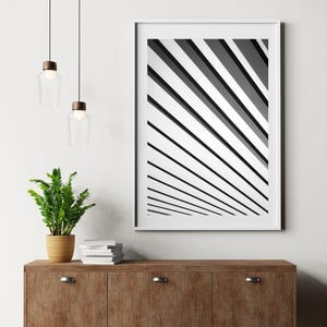 Può includere: Una stampa artistica astratta in bianco e nero incorniciata con linee diagonali. L'opera d'arte è esposta su un mobile in legno con una pianta e lampade a sospensione.
