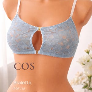 Puede incluir: Un bralette de encaje azul claro con tirantes blancos y un cierre dorado. El bralette tiene un diseño de ojo de cerradura en el centro. La palabra "COS" está impresa encima de la palabra "Bralette".