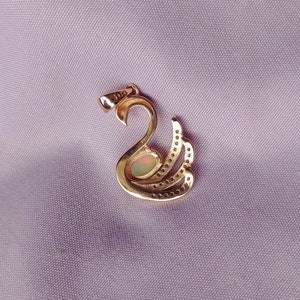 May include: Gold swan pendant with a white opal gemstone in the center. The swan has a detailed design with small stones around the edges.