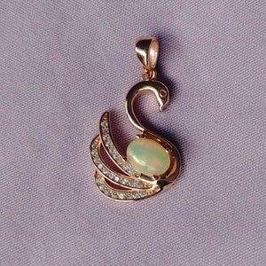 May include: A rose gold pendant featuring a swan design with a clear opal gemstone in the center. The swan's wings are adorned with small, clear cubic zirconia stones.