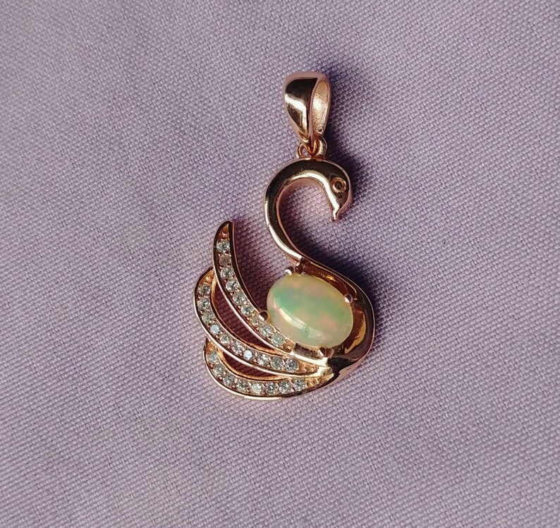 May include: A rose gold pendant featuring a swan design with a clear opal gemstone in the center. The swan's wings are adorned with small, clear cubic zirconia stones.