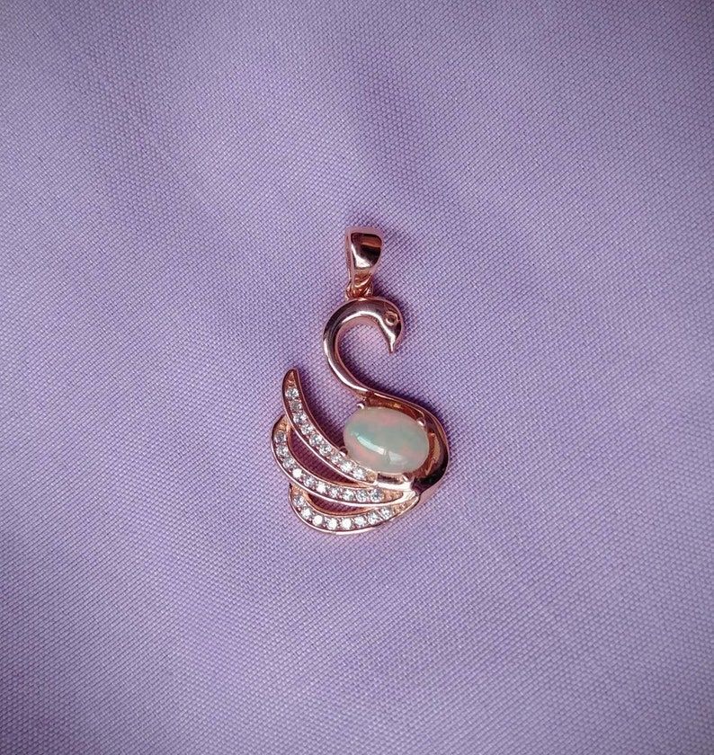 May include: A rose gold swan pendant with a white opal gemstone in the center. The swan is adorned with small clear cubic zirconia stones.