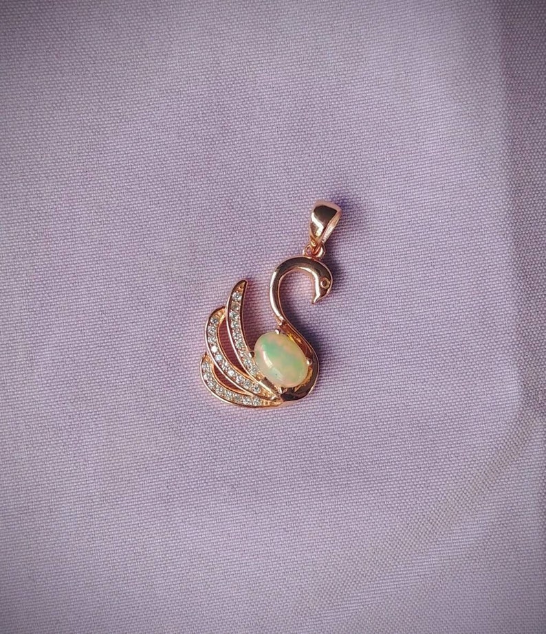 May include: A rose gold pendant featuring a swan design with a clear opal gemstone in the center. The swan's wings are adorned with small clear cubic zirconia stones.