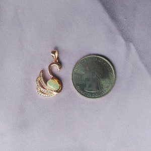 May include: A rose gold swan pendant with a white opal stone and clear cubic zirconia accents. The pendant is shown next to a US quarter for size comparison.