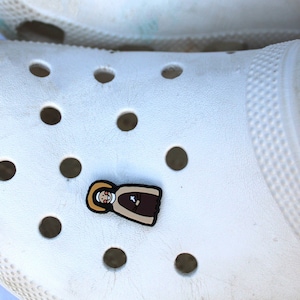 Könnte beinhalten: Weißer Clog mit einem braunen und gelben Cartoon-Charakter-Anhänger, der an der Vorderseite befestigt ist. Der Anhänger ist eine Cartoon-Darstellung einer Person, die eine braune Robe und einen gelben Heiligenschein trägt.