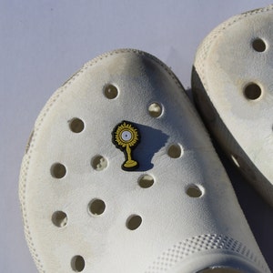 May include: A white pair of Crocs with a gold and black religious pin featuring a sunburst and a cross. The pin is attached to one of the shoes.