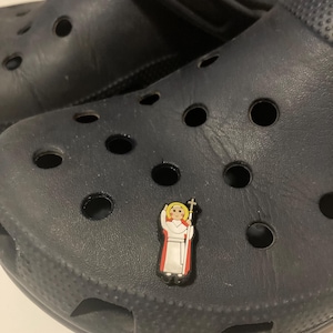 May include: A blue pair of clogs with a small, colorful charm of a person in a white robe and red sash holding a staff.