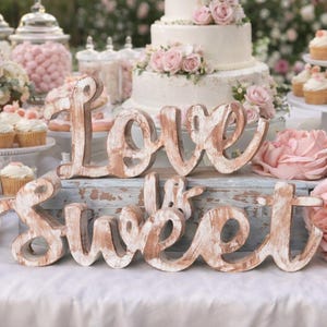May include: A dessert table with a three-tiered cake adorned with pink roses, cupcakes, donuts, and the wooden words "Love is Sweet." The table is decorated with pink flowers and candies, creating a romantic and celebratory atmosphere.