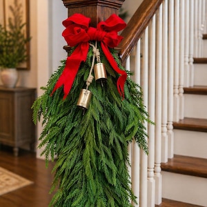 May include: A festive Christmas garland made of evergreen branches, adorned with a large red bow and two gold bells. The garland is draped over a wooden staircase post, creating a welcoming holiday decoration.