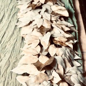 May include: A garland of faux leaves in shades of beige and green. The leaves are arranged in a cascading pattern, creating a lush and natural look.
