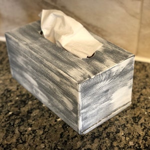 May include: A gray wooden tissue box with a distressed finish. The box has a rectangular shape and a slot for tissues on the top.