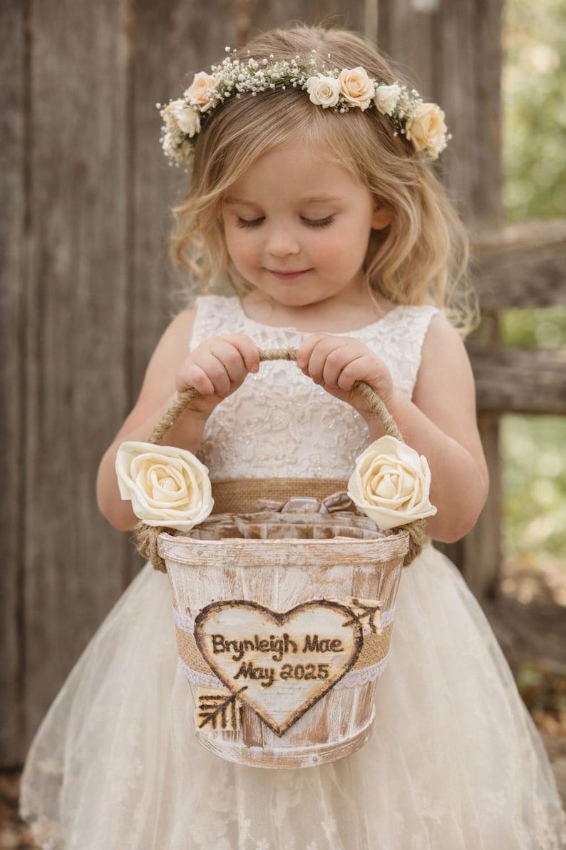 May include: A young girl wearing a white dress and a floral crown holds a rustic flower basket. The basket is decorated with cream-colored roses and a heart-shaped emblem with the name "Brynleigh Mae" and the date "May 2025".