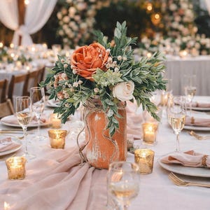 May include: A rustic centerpiece featuring an orange-colored flower arrangement in a distressed jar, accented with greenery and twine. The table setting includes champagne glasses, candles, and a blush-colored runner, creating an elegant ambiance for a special event.