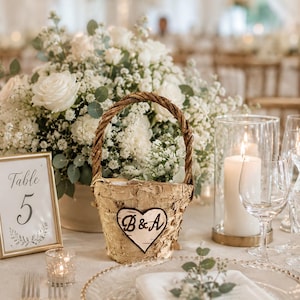 May include: A wedding table setting with a birch bark basket featuring a heart with "B&A" initials, surrounded by white roses and baby's breath. A framed table number 5, candles, and glassware complete the elegant decor.