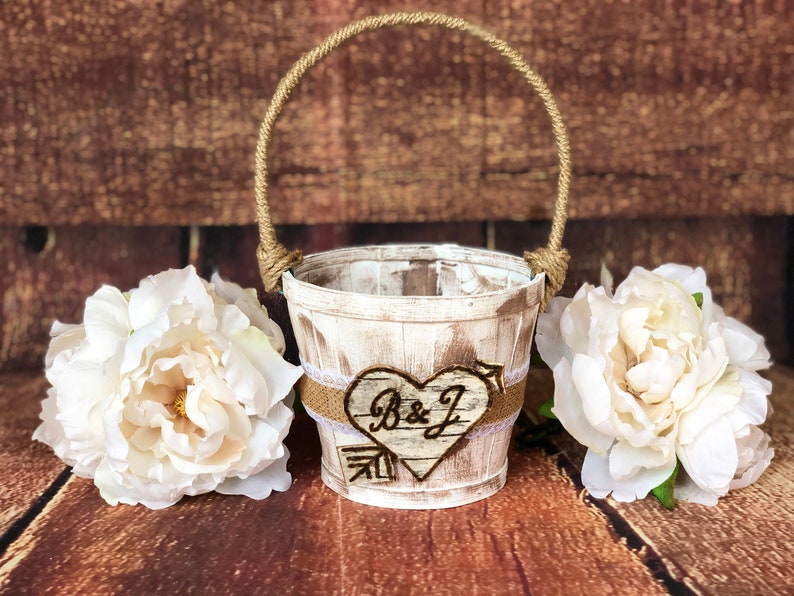 May include: A rustic, white-washed wooden flower girl basket with a jute rope handle. The basket features a heart-shaped wooden accent with the initials "B & J". Two large white peony flowers flank the basket.