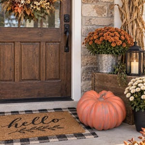 May include: A rectangular coir doormat with the word hello and a leaf branch graphic printed in black, layered over a black and white buffalo check rug. The scene includes a large orange pumpkin, potted orange mums, and a fall wreath.
