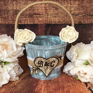 May include: A rustic, distressed blue wooden bucket with a rope handle, adorned with cream-colored fabric roses. A burlap and lace band wraps the bucket, featuring a wooden heart with the initials "B & C".