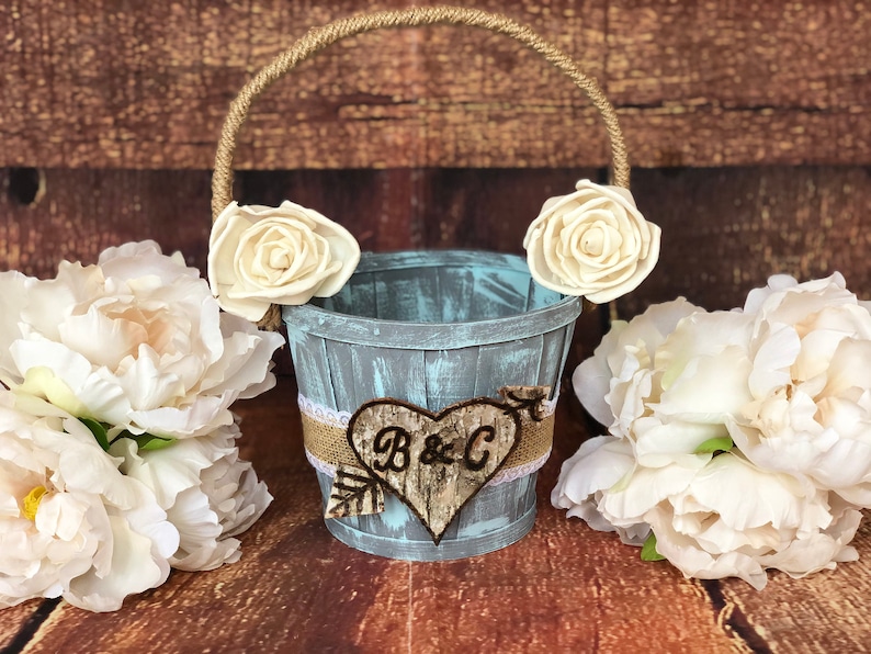 May include: A rustic, distressed blue wooden bucket with a rope handle, adorned with cream-colored fabric roses. A burlap and lace band wraps the bucket, featuring a wooden heart with the initials "B & C".