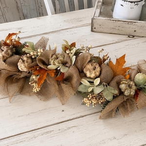 May include: A decorative table runner featuring tan burlap fabric loops, artificial pumpkins in muted tones, orange and brown maple leaves, and clusters of small white berries. The arrangement is designed for horizontal display on a flat surface.