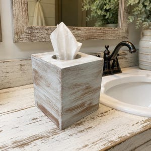 May include: A square tissue box cover featuring a distressed finish with visible white paint over a natural brown wood grain. The box has a circular opening at the top with a single white facial tissue protruding from the center of the container.