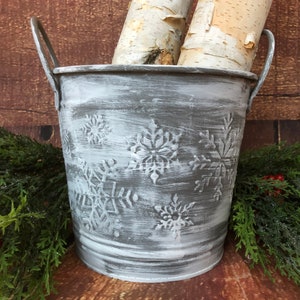 May include: A white metal bucket with snowflake designs, distressed with a gray wash. The bucket has two handles and is filled with birch logs.
