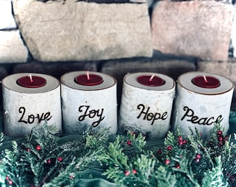 May include: Four birch wood candle holders with red candles, each inscribed with a word: Love, Joy, Hope, and Peace. The candle holders are arranged in a row, with greenery and red berries in the foreground, against a stone background.
