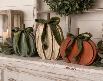 May include: Three wooden pumpkin decorations in teal, cream, and orange, each featuring a distressed finish, a brown stem, and a dark green frayed fabric ribbon tied in a bow around the stem, arranged in a row on a white surface.