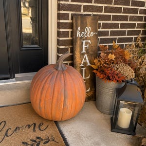 May include: A large orange pumpkin with a brown stem, a tall rectangular wooden sign with the white text hello FALL, a galvanized metal bucket filled with orange and brown autumn foliage, and a black lantern containing a white pillar candle.