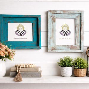 May include: Two framed prints with a rustic, distressed look hang on a white wall. One frame is teal, the other is light blue. The prints feature a logo with a barn and the text "The Little Rustic Farm". A vase of flowers and other decor sit below.