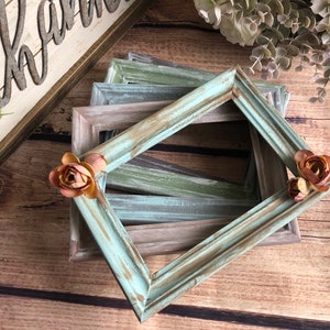 May include: A set of four rustic wooden picture frames in shades of brown and blue-green. The frames are stacked and distressed, giving them a vintage look. Two small faux flowers are placed on the top frame.