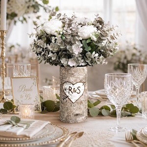 May include: A wedding table setting with a birch bark vase holding white flowers and greenery. The vase has a heart with the initials "R&S". Crystal wine glasses, candles, and a "Bride to Be" sign complete the elegant decor.