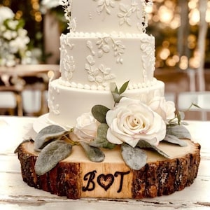 May include: A three-tiered white wedding cake with floral decorations. The cake sits on a rustic wooden base adorned with white roses and greenery. The initials "B♡T" are carved into the wood.