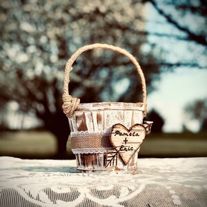 May include: A small, distressed white wooden basket with a jute handle and a burlap and lace band. A wooden heart with "Pamela + Eric" is attached. The basket sits on a white lace tablecloth.