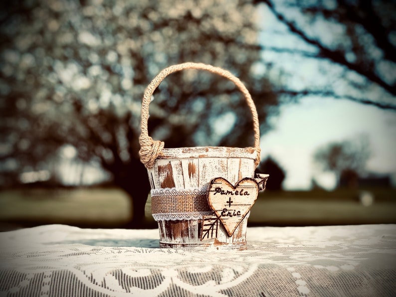 May include: A small, distressed white wooden basket with a jute handle and a burlap and lace band. A wooden heart with "Pamela + Eric" is attached. The basket sits on a white lace tablecloth.