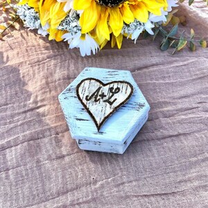 May include: A white, hexagonal wooden box with a distressed finish. A heart-shaped design with the initials "A & L" is burned into the wood. The box is on a textured, light brown surface, with yellow and white flowers in the background.