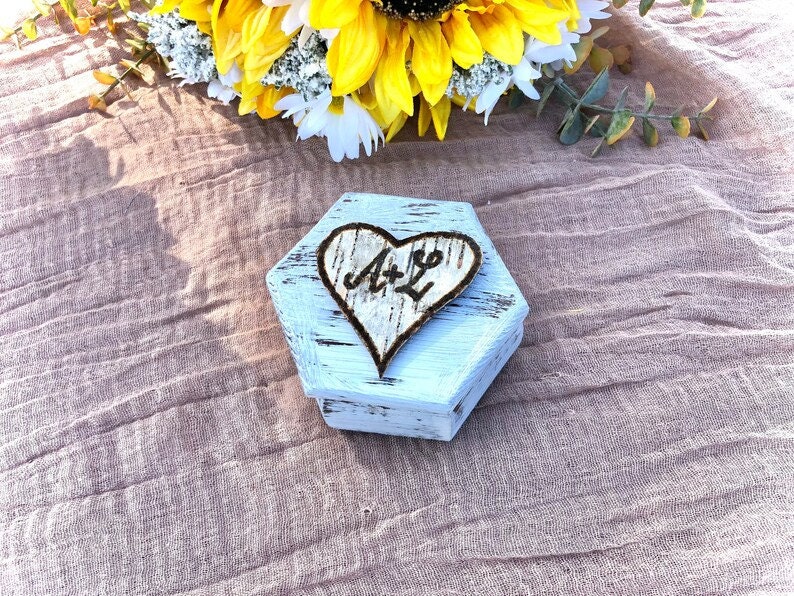 May include: A white, hexagonal wooden box with a distressed finish. A heart-shaped design with the initials "A & L" is burned into the wood. The box is on a textured, light brown surface, with yellow and white flowers in the background.
