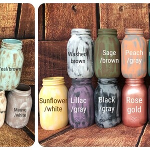 May include: A collection of painted mason jars in various colors, including dark gray, navy, pink, teal, and sunflower. Each jar has a distressed, rustic finish. The jars are labeled with color combinations like "Washed brown" and "Rose gold."