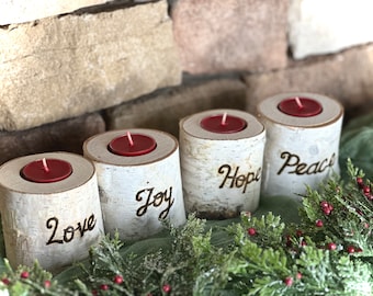 May include: Four birch log candle holders with red candles. Each holder has a word burned into the wood: Love, Joy, Hope, and Peace. The candles sit on a green cloth with greenery and red berries.