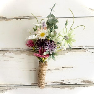 May include: A floral boutonniere with a variety of colorful flowers, including daisies, purple alliums, and pink blooms. The stems are wrapped in twine, creating a rustic and natural look. The arrangement is set against a white, weathered wooden background.