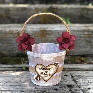 May include: A rustic wedding basket with a jute handle and burgundy flower accents. The basket features a heart-shaped wooden emblem with the names "Amber & Dan" and an arrow design, perfect for a wedding ceremony.