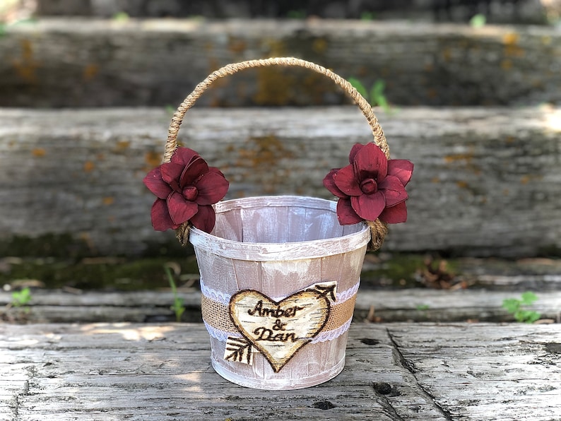 May include: A rustic wedding basket with a jute handle and burgundy flower accents. The basket features a heart-shaped wooden emblem with the names "Amber & Dan" and an arrow design, perfect for a wedding ceremony.