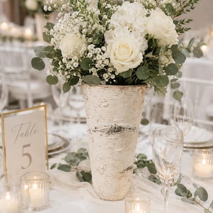 May include: A tall birch bark vase overflowing with white roses, hydrangeas, and baby's breath, accented with green foliage. The vase sits on a table set for an event, with candles and a table number 5 sign.
