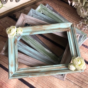 May include: A stack of four rustic wooden picture frames in various shades of blue, green, and brown. The frames are distressed and have a vintage look. Each frame is adorned with a white flower.