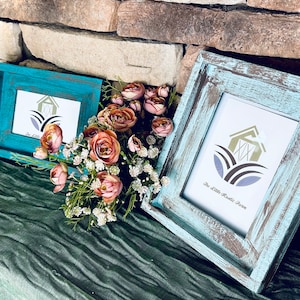 May include: Two rustic, distressed wooden picture frames in teal and light blue, each holding a print with a barn logo and the text "The Little Rustic Farm." A bouquet of pink and white flowers sits between the frames.